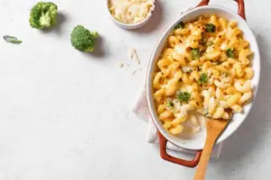 The image shows a dish of creamy macaroni and cheese, garnished with small pieces of broccoli. The dish is served in an oval ceramic baking dish with a wooden spoon placed inside. In the background, there are some broccoli florets and a small bowl containing grated cheese. The setting is clean and minimalistic, with a light-colored surface.





