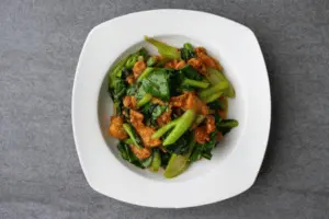 The image shows a dish of stir-fried vegetables and meat, likely pork or chicken, served on a white plate. The vegetables appear to include bok choy or another type of leafy green, along with other green stalks. The meat is coated in a savory sauce, giving it a slightly glazed appearance. The dish is plated simply, with no additional garnishes, on a dark grey background.