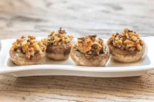 The image shows a dish of stuffed mushrooms arranged on a rectangular white platter. Each mushroom cap is filled with a savory stuffing that appears to include breadcrumbs, herbs, and possibly cheese or minced vegetables. The mushrooms are baked or roasted, giving the stuffing a golden-brown, crispy texture on top. The setting is rustic, with a wooden table beneath the plate.






